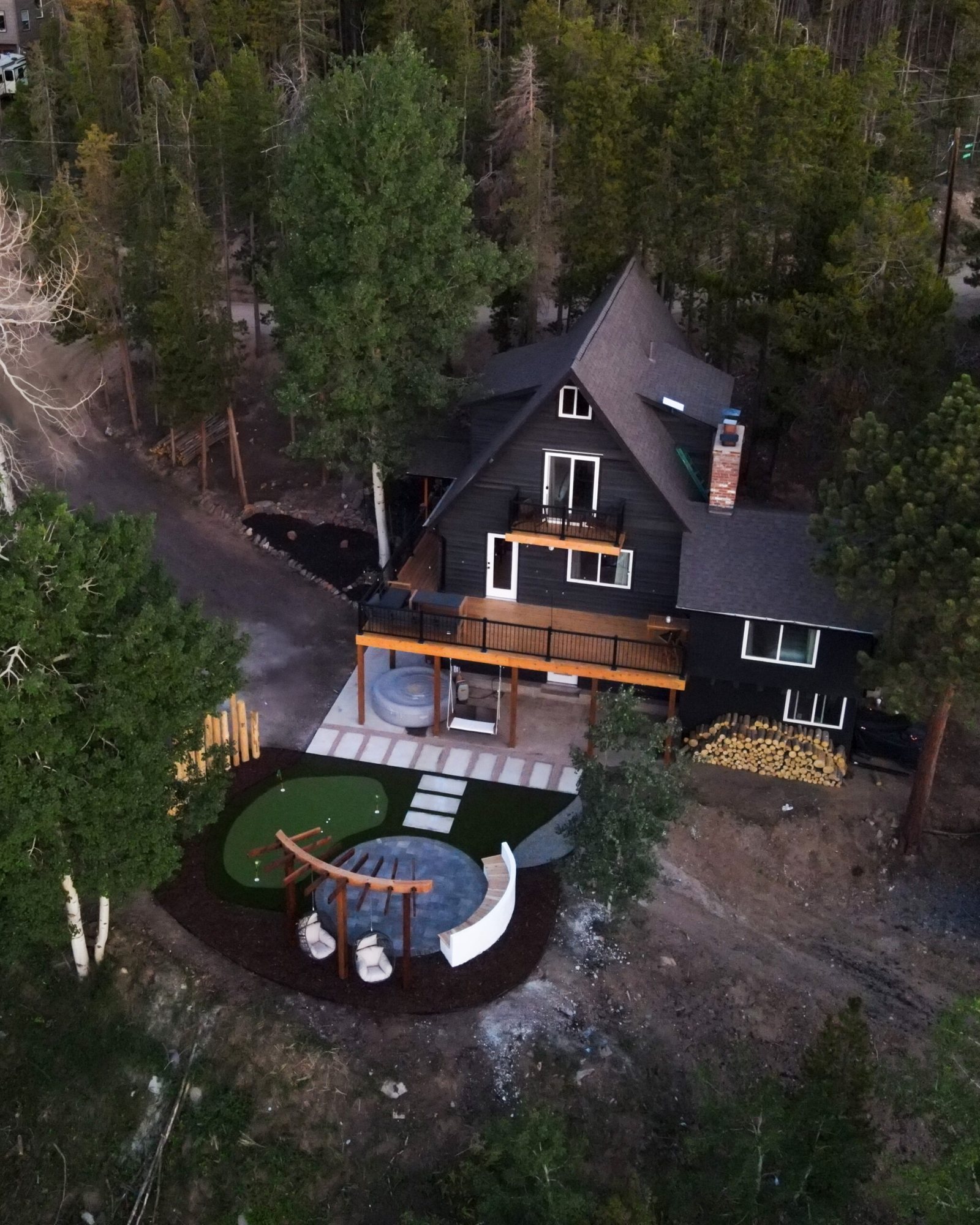 Aerial view of an A-frame house with a lower patio, pergola and putting green among trees