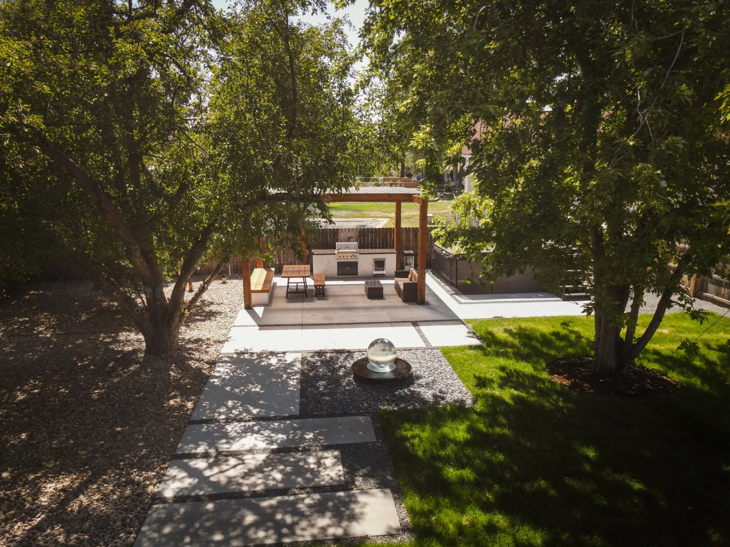 Backyard with a covered outdoor kitchen and pergola, paver patio and lawn under mature trees